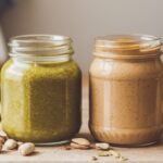 Jars of pistachio butter and peanut butter side by side with nuts scattered around.