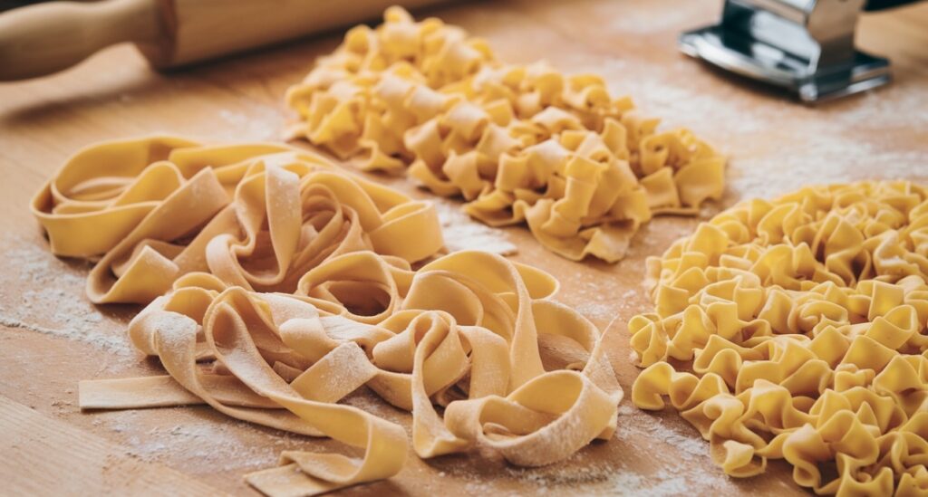 "Freshly made pappardelle ribbons and egg noodles on a wooden surface, showing their shape and texture differences