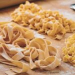 "Freshly made pappardelle ribbons and egg noodles on a wooden surface, showing their shape and texture differences